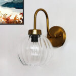 Brass wall sconce with curved arm and clear ribbed glass globe shade mounted on a white wall, featuring an exposed bulb for bright ambient lighting.