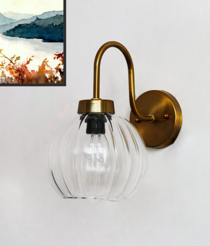 Brass wall sconce with curved arm and clear ribbed glass globe shade mounted on a white wall, featuring an exposed bulb for bright ambient lighting.