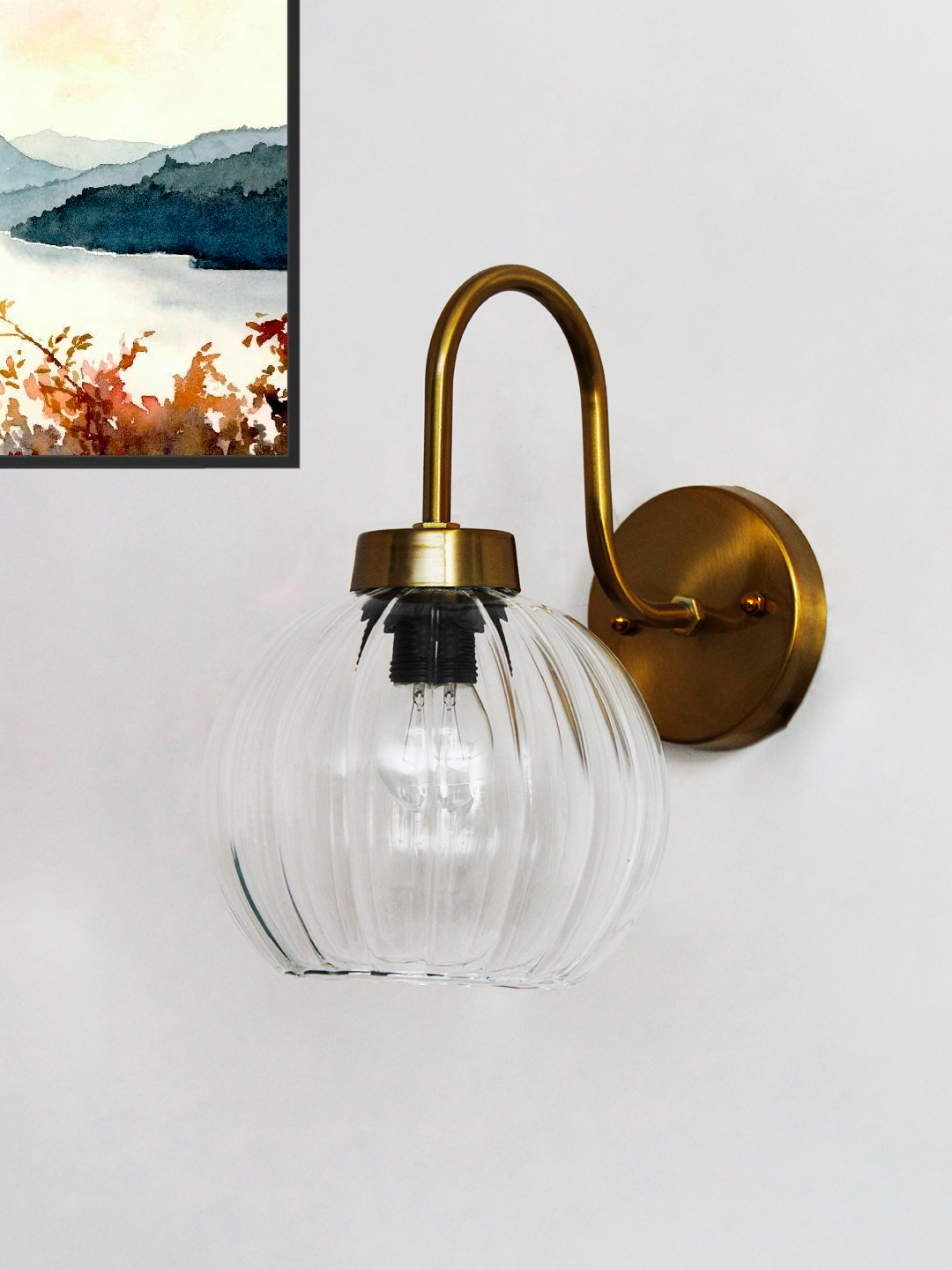 Modern Brass Wall Sconce Clear Ribbed Glass Light Brass wall sconce with curved arm and clear ribbed glass globe shade mounted on a white wall, featuring an exposed bulb for bright ambient lighting.