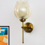 Modern wall sconce with amber glass shade and gold metal base, providing warm ambient lighting on a clean minimalist interior wall.