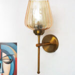 Wall-mounted brass sconce with ribbed glass shade casting warm ambient light beside a colorful abstract portrait on a white wall