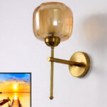 Modern light fixture wall sconce with amber glass shade and brass finish, casting warm light beside a sunset landscape wall art on a white wall
