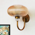 Brass wall sconce with curved metal arm and amber ribbed glass shade mounted on a white wall, casting warm ambient light in a modern interior.