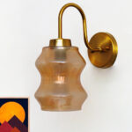 Vintage brass wall sconce with curved arm and amber glass shade mounted on a white wall, casting warm ambient light.