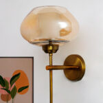 Big Kachori Glass wall light with amber dome-shaped glass shade and brass metal mount installed on a white wall, creating a warm ambient glow beside a framed artwork