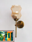 Damru Glass wall light with hourglass-shaped amber glass shade and brass metal mount on a white wall, casting a warm ambient glow beside a colorful framed artwork