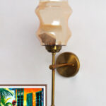 Damru Glass wall light with hourglass-shaped amber glass shade and brass metal mount on a white wall, casting a warm ambient glow beside a colorful framed artwork