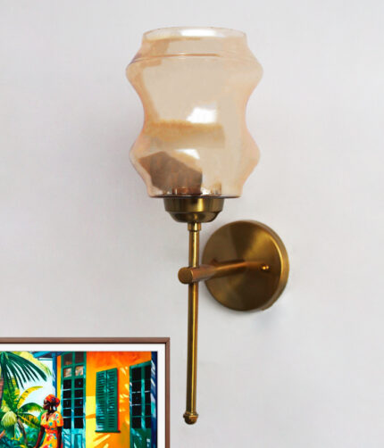 Damru Glass wall light with hourglass-shaped amber glass shade and brass metal mount on a white wall, casting a warm ambient glow beside a colorful framed artwork