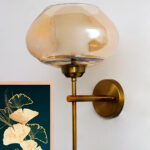 Small Kachori Glass wall light featuring a rounded amber glass shade with a brass metal mount, installed on a white wall beside a decorative floral artwork, emitting warm ambient lighting.
