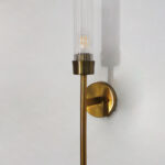 Slim linear Wall Lamp with brass finish and vertical cylindrical glass shade mounted on a wall, featuring a minimalist modern design and warm ambient lighting.