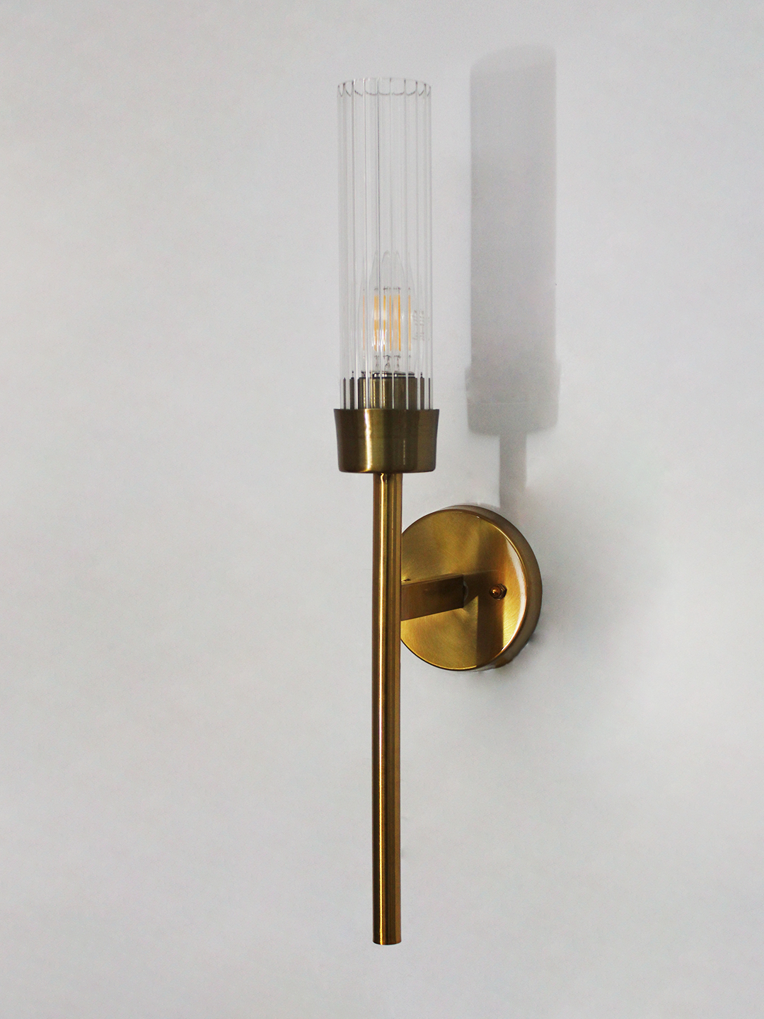 Clear Glass Slim linear Wall Lamp Brass Slim linear Wall Lamp with brass finish and vertical cylindrical glass shade mounted on a wall, featuring a minimalist modern design and warm ambient lighting.
