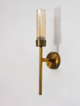 Linear wall lamp in brass finish with a slim vertical rod and ribbed amber glass cylindrical shade, mounted on a wall for modern decorative lighting