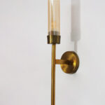 Linear wall lamp in brass finish with a slim vertical rod and ribbed amber glass cylindrical shade, mounted on a wall for modern decorative lighting