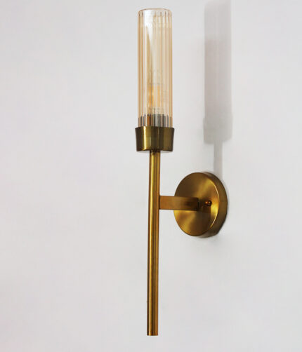 Linear wall lamp in brass finish with a slim vertical rod and ribbed amber glass cylindrical shade, mounted on a wall for modern decorative lighting