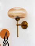 Modern brushed brass wall sconce with ribbed amber glass shade, mounted on a white wall, providing warm ambient lighting for contemporary interiors