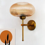 Modern brushed brass wall sconce with ribbed amber glass shade, mounted on a white wall, providing warm ambient lighting for contemporary interiors