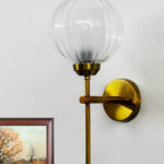 Clear Glass Wall Light with a rounded transparent glass shade and brushed brass arm, mounted on a white wall, providing soft ambient lighting in a modern interior setting