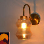 Vintage brass wall sconce with curved arm and amber glass shade mounted on a white wall, casting warm ambient light.