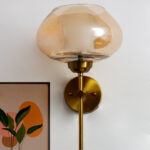 Big Kachori Glass wall light with amber dome-shaped glass shade and brass metal mount installed on a white wall, creating a warm ambient glow beside a framed artwork