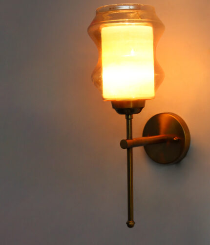 Damru Glass wall light with hourglass-shaped amber glass shade and brass metal mount on a white wall, casting a warm ambient glow beside a colorful framed artwork