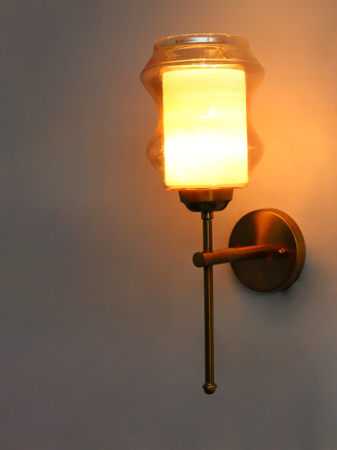 Damru Glass Amber Wall Sconce Brass Lighting Damru Glass wall light with hourglass-shaped amber glass shade and brass metal mount on a white wall, casting a warm ambient glow beside a colorful framed artwork
