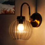 Brass wall sconce with curved arm and clear ribbed glass globe shade mounted on a white wall, featuring an exposed bulb for bright ambient lighting.