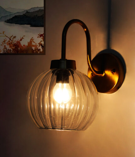 Brass wall sconce with curved arm and clear ribbed glass globe shade mounted on a white wall, featuring an exposed bulb for bright ambient lighting.