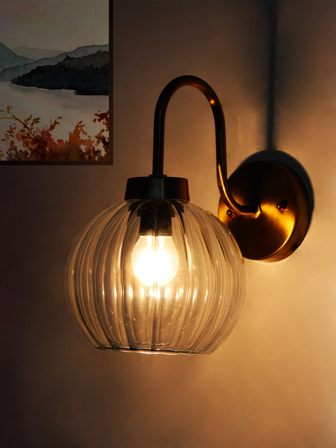 Modern Brass Wall Sconce Clear Ribbed Glass Light Brass wall sconce with curved arm and clear ribbed glass globe shade mounted on a white wall, featuring an exposed bulb for bright ambient lighting.
