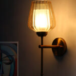 Wall-mounted brass sconce with ribbed glass shade casting warm ambient light beside a colorful abstract portrait on a white wall