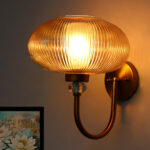 Brass wall sconce with curved metal arm and amber ribbed glass shade mounted on a white wall, casting warm ambient light in a modern interior.