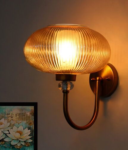 Brass wall sconce with curved metal arm and amber ribbed glass shade mounted on a white wall, casting warm ambient light in a modern interior.