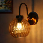 Brass wall sconce with curved arm and ribbed amber glass globe shade mounted on a white wall, providing warm ambient lighting.