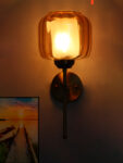 Modern light fixture wall sconce with amber glass shade and brass finish, casting warm light beside a sunset landscape wall art on a white wall