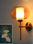 Small Kachori Glass wall light featuring a rounded amber glass shade with a brass metal mount, installed on a white wall beside a decorative floral artwork, emitting warm ambient lighting.