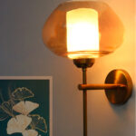 Small Kachori Glass wall light featuring a rounded amber glass shade with a brass metal mount, installed on a white wall beside a decorative floral artwork, emitting warm ambient lighting.