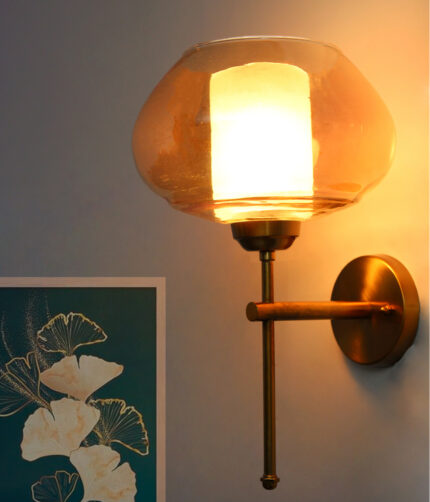 Small Kachori Glass wall light featuring a rounded amber glass shade with a brass metal mount, installed on a white wall beside a decorative floral artwork, emitting warm ambient lighting.