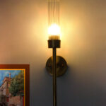 Slim Tube Wall Lamp with brass finish and vertical cylindrical glass shade mounted on a wall, featuring a minimalist modern design and warm ambient lighting.
