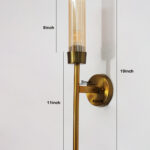 Linear wall lamp in brass finish with a slim vertical rod and ribbed amber glass cylindrical shade, mounted on a wall for modern decorative lighting