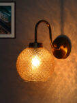 Modern wall sconce with amber textured glass globe shade and brushed brass finish, emitting warm ambient light on wall, ideal for bedroom, hallway, or living room décor.