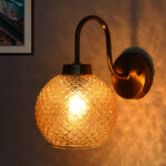 Modern wall sconce with amber textured glass globe shade and brushed brass finish, emitting warm ambient light on wall, ideal for bedroom, hallway, or living room décor.