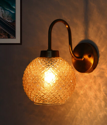 Modern wall sconce with amber textured glass globe shade and brushed brass finish, emitting warm ambient light on wall, ideal for bedroom, hallway, or living room décor.